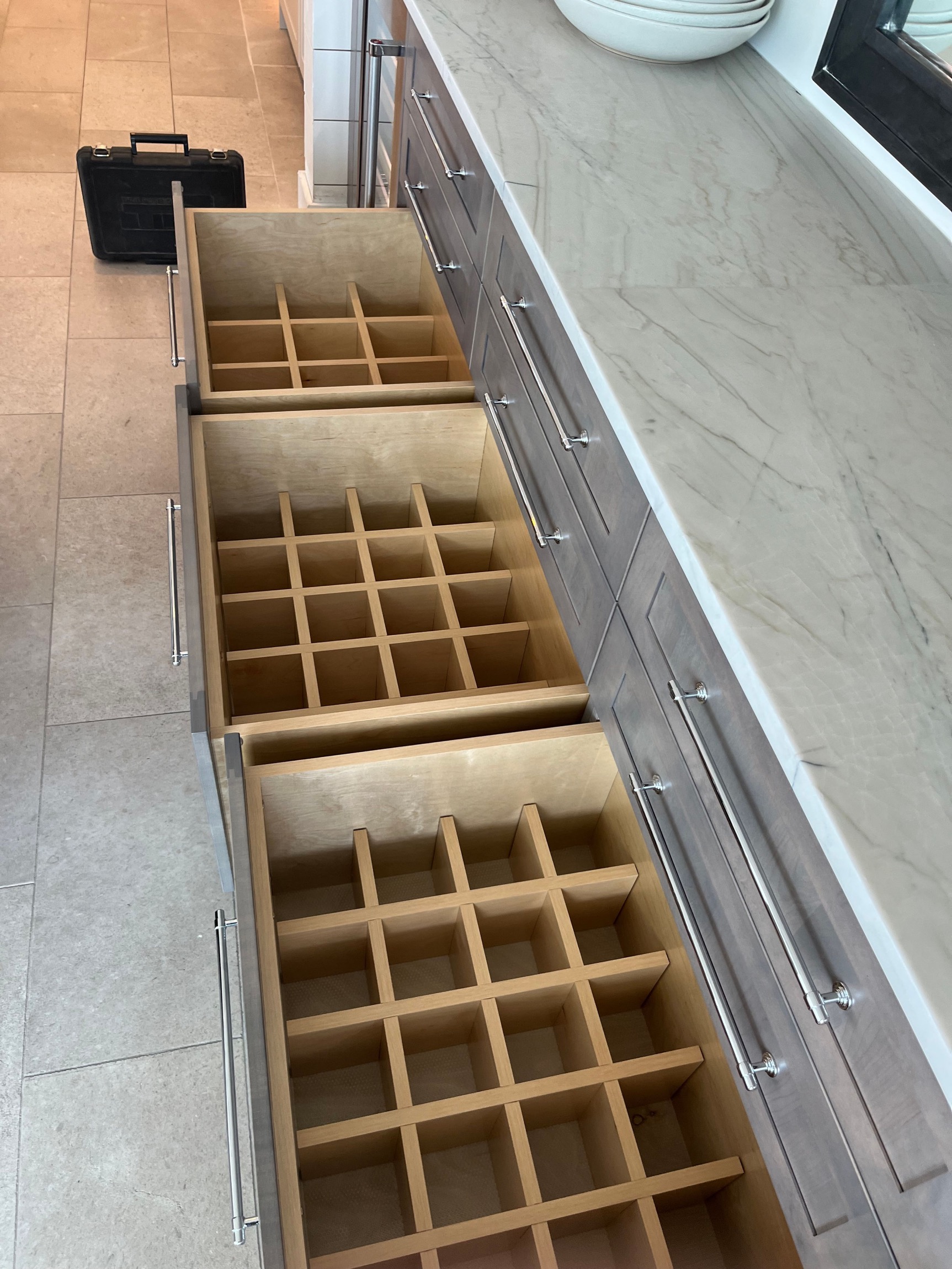 Open kitchen drawers with grid dividers for storing bottles or glasses, beside marble countertop.
