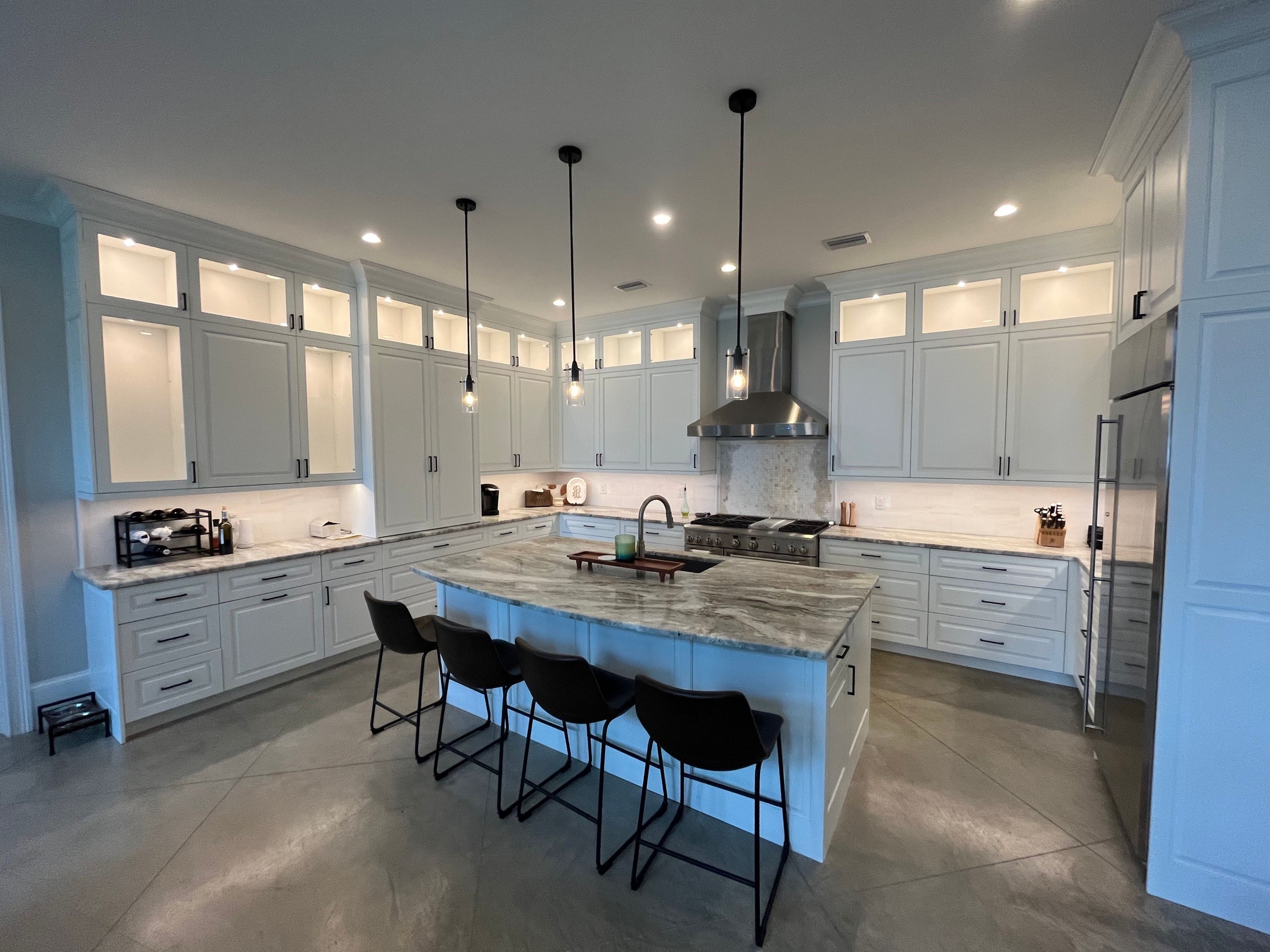 White kitchen with marble island, black bar stools, pendant lights, stainless appliances, and polished concrete floor.