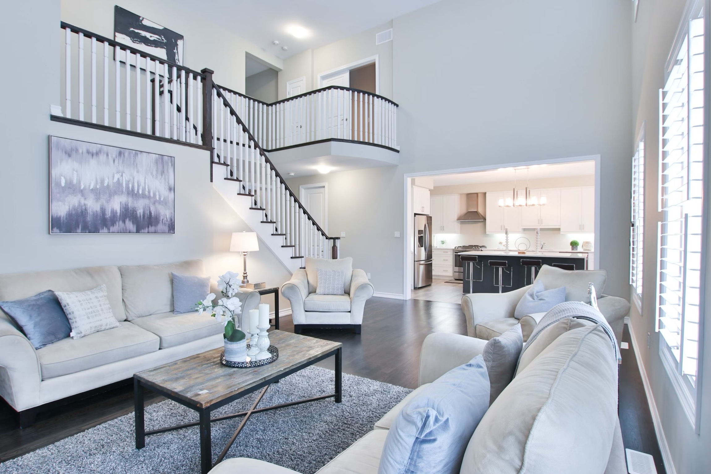Two-story living room with white sofas, coffee table, staircase, and open view to kitchen with island.