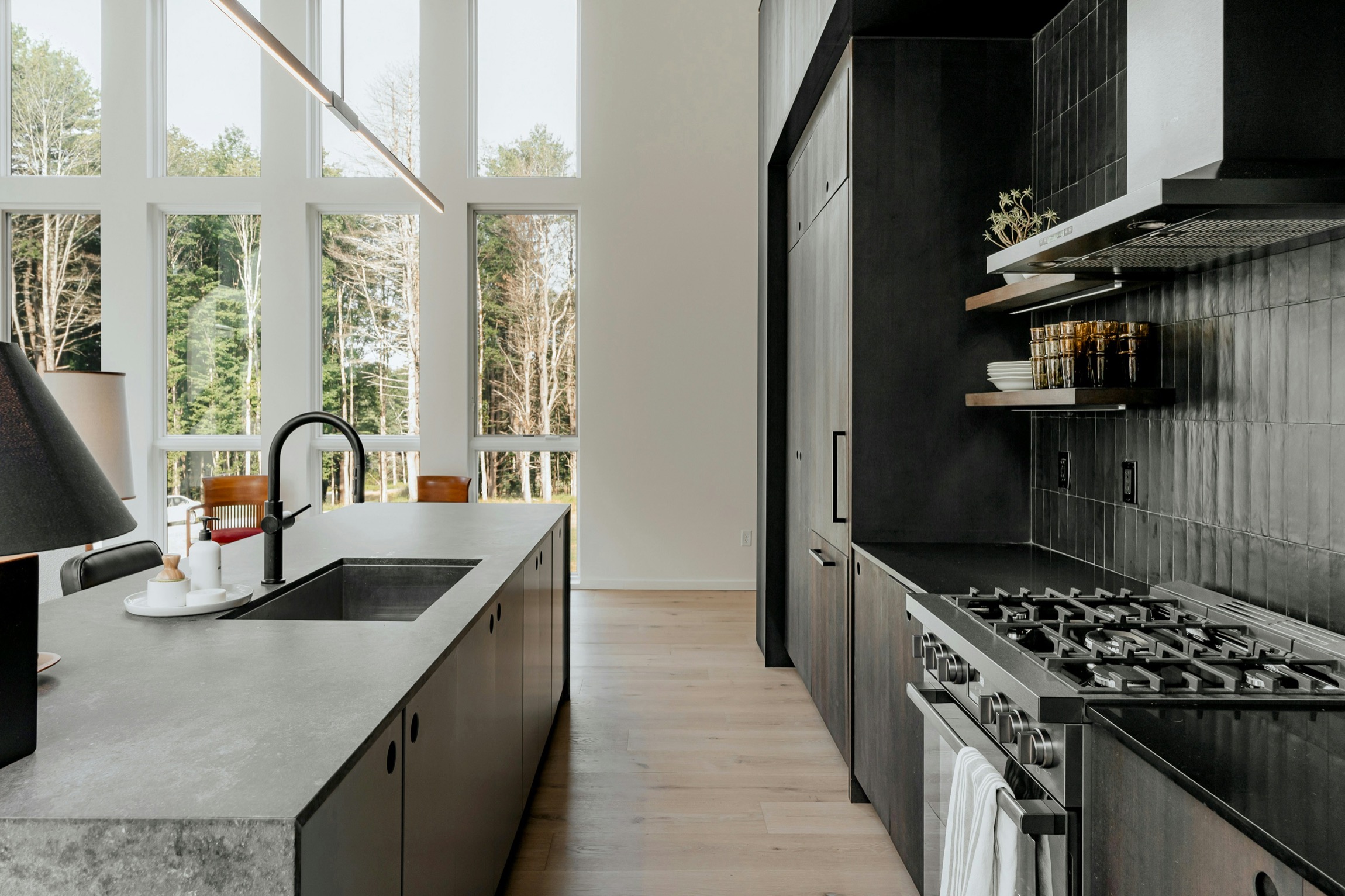 Modern kitchen with dark cabinets, stainless stove, island with sink, and large windows overlooking trees.