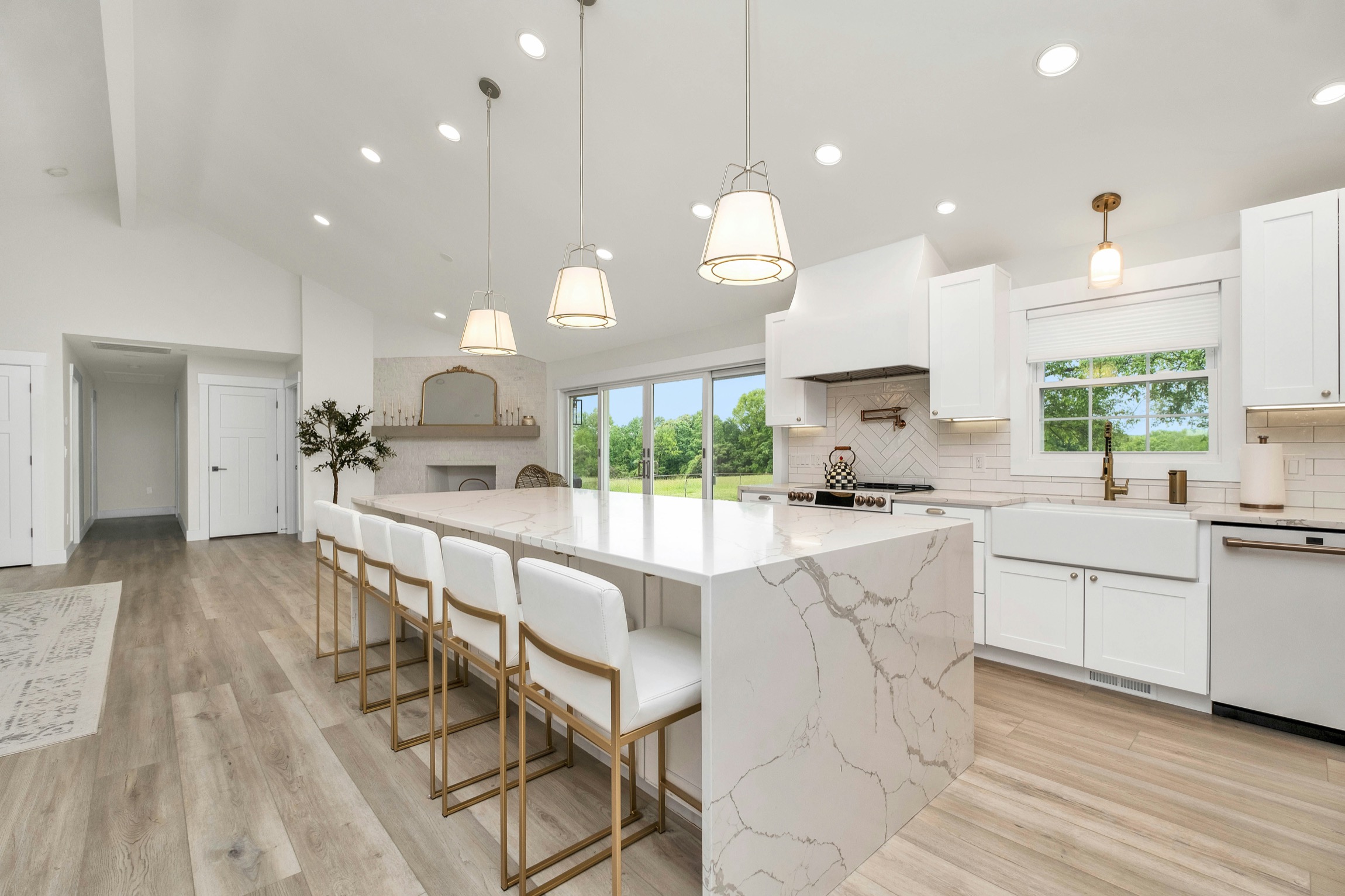 Modern white kitchen with marble island, gold-legged bar stools, pendant lights, and vaulted ceiling.