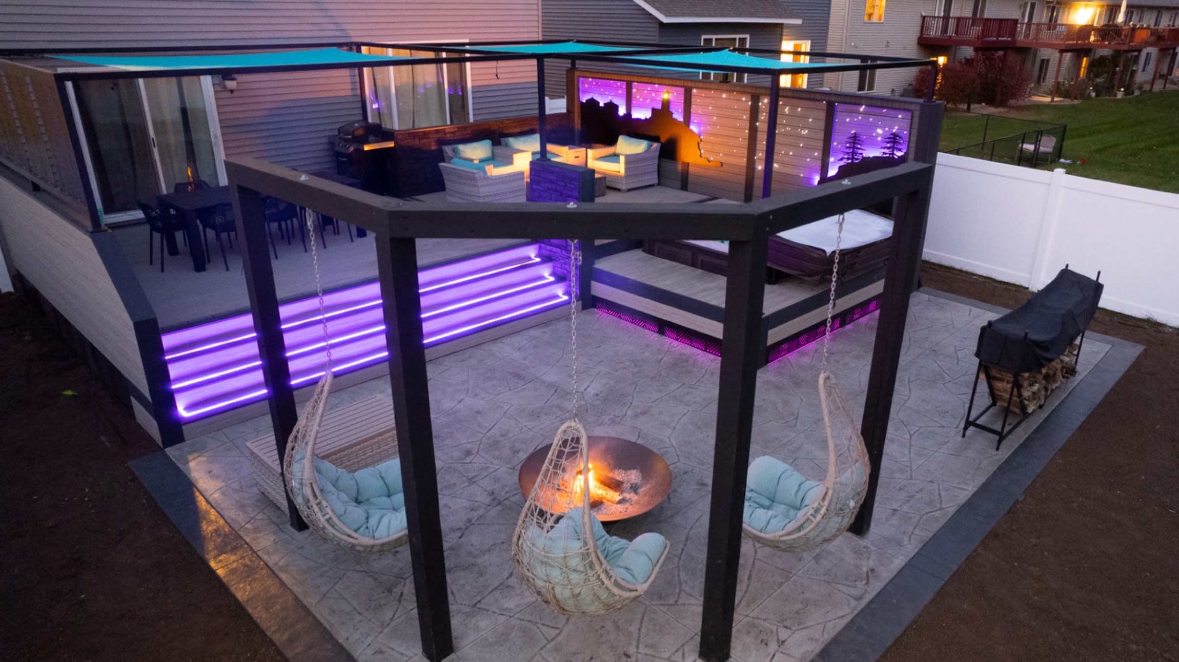Two-level backyard deck lit with purple LED lighting at dusk with hanging chairs and a fire pit below