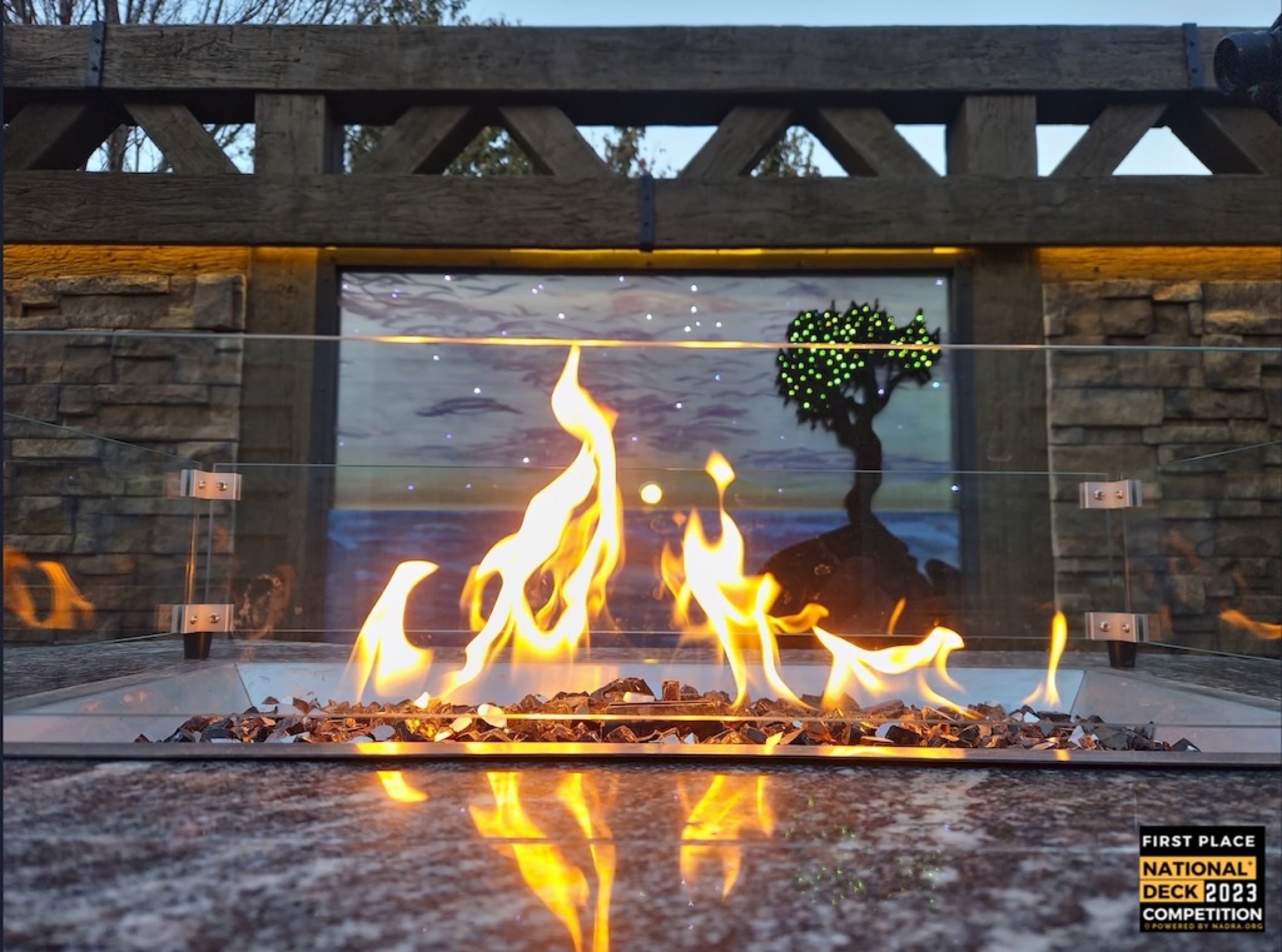 Glass-enclosed fire feature with tall flames burning over lava rock in front of a stained-glass tree panel and stone wall.