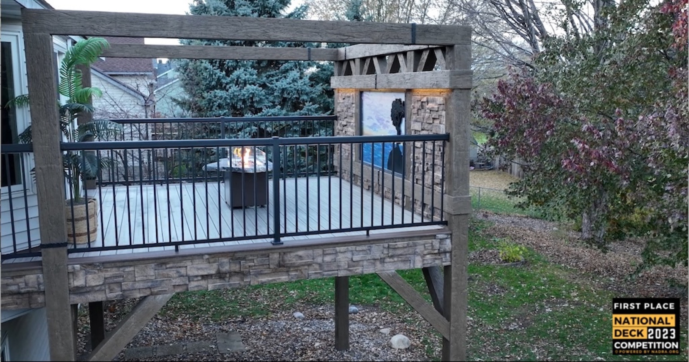 Elevated deck with stone base, black metal railing, pergola, lit fire table, and stone accent wall in a backyard.